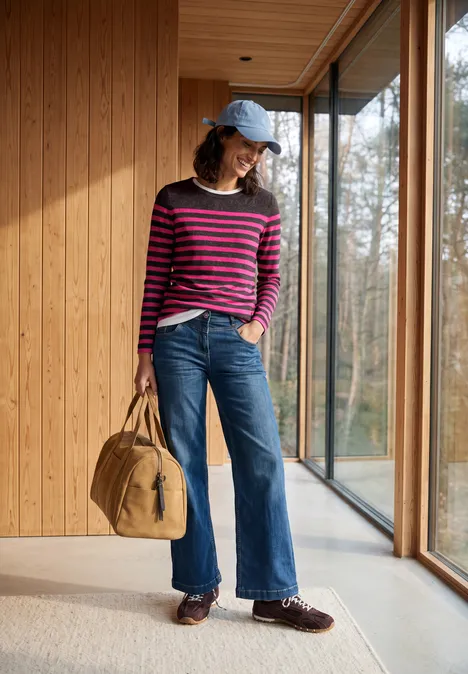 Vrouw in roze-blauw gestreepte trui, wijd jeans, blauwe pet en tas staat in een lichte houten gang met ramen.
