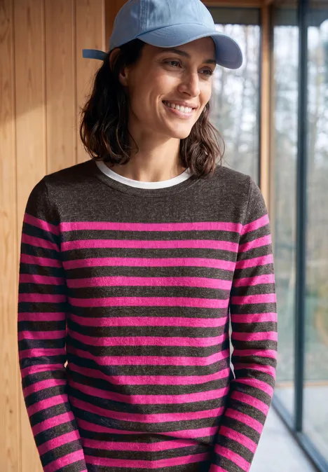 Vrouw met blauwe pet en grijs-fuchsia gestreepte trui, glimlachend voor een raam.