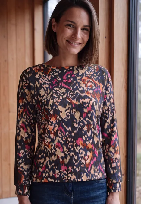 Een glimlachende vrouw staat bij een raam in een kleurrijk, patroonshirt met lange mouwen.