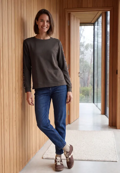 Shirt mit Boat Neck und Print-Tape truffle brown Eine Frau in braunem Pullover und Jeans steht lächelnd an einer Holzverkleidung vor großen Fenstern.