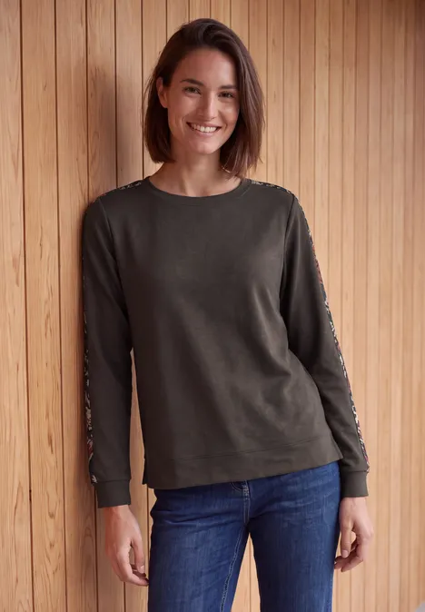Shirt mit Boat Neck und Print-Tape truffle brown Eine lächelnde Frau mit schulterlangem Haar, grauem Langarmshirt und blauer Jeans vor Holzwand.