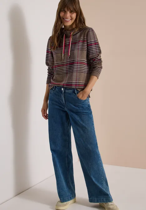 Langarm Shirt mit Funnel Neck und Muster dark toffee brown Frau mit kariertem Kapuzenpullover und weiten Jeans vor neutralem Hintergrund lächelnd.