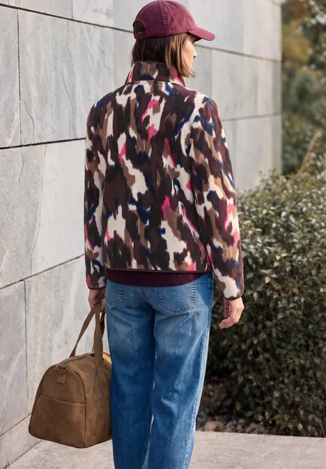 Vrouw met rode pet, dessinjas en blauwe jeans draagt een bruine tas voor een muur en struiken.
