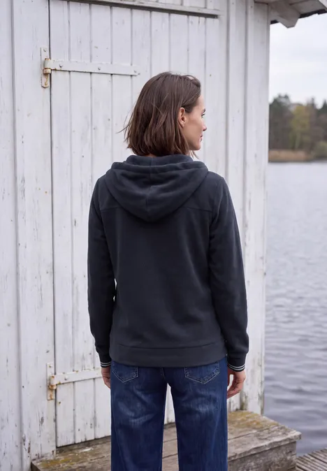 Rückansicht einer Person in dunkelgrauer Kapuzenjacke vor einem weißen Holzhaus am Wasser.