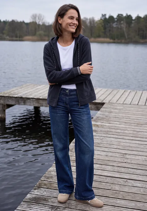 Frau lächelnd in Jeans und Cardigan steht auf einem Steg am ruhigen See mit Wald im Hintergrund.