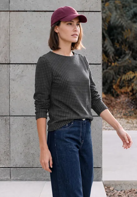 Frau mit rosafarbenem Cap, grauem Pullover und Jeans steht vor Wand und Pflanzen.