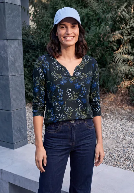 Eine Frau mit weißer Kappe, blau-grün gemustertem Oberteil und Jeans steht draußen und lächelt.