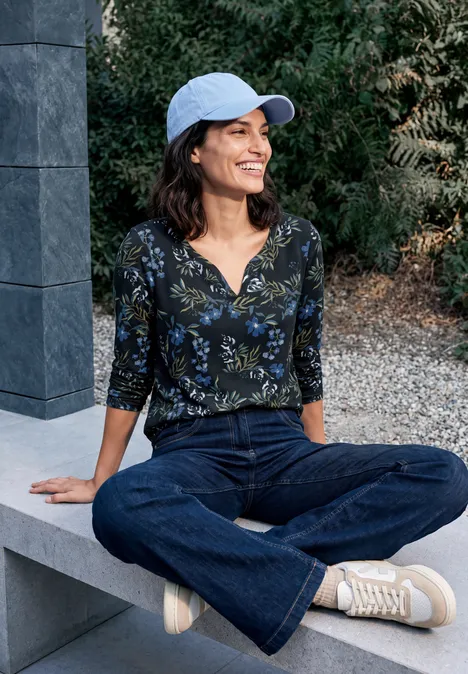 Frau mit blauer Cap sitzt lächelnd im Schneidersitz auf Steinbank, trägt Blumenoberteil, Jeans und weiße Sneaker.