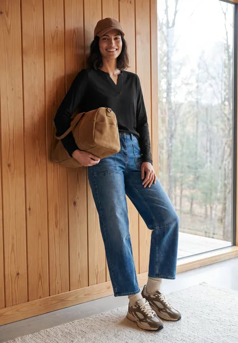 Frau in Jeans, dunkler Jacke und Cap steht mit Beuteltasche lächelnd an Holzwand neben Fenster.