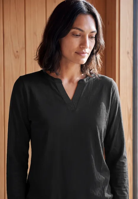 Eine Frau in schwarzem Langarmshirt steht vor Holzpaneelen und blickt nachdenklich zur Seite.