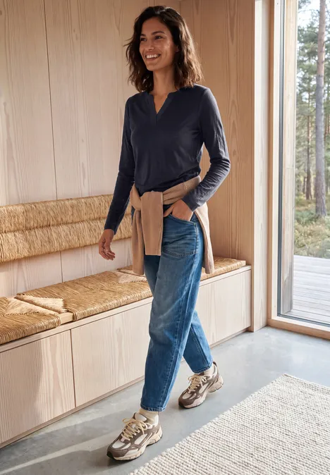 Frau in Jeans, dunkelblauem Shirt und Sneakern geht in modernem, hellem Raum an einer Holzbank vorbei.