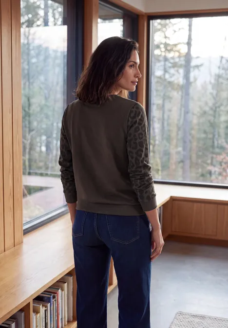 Shirt met ronde hals en luipaardprint op de mouwen truffle brown Frau steht rücklings in einem hellen Holzraum, lehnt an Sitzbank, trägt Jeans und grünen Pullover.