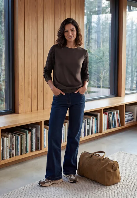Shirt met ronde hals en luipaardprint op de mouwen truffle brown Eine Frau in Jeans, Pullover und Sneakers steht in einem hellen Bücherraum mit Reisetasche.