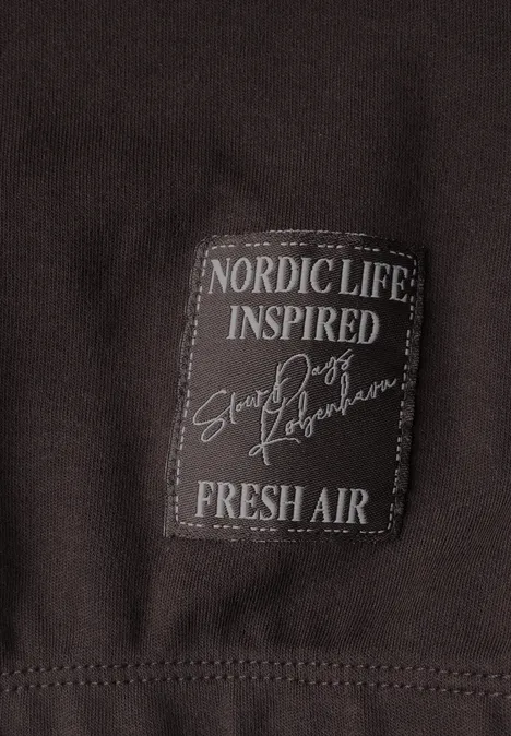 Shirt met ronde hals en luipaardprint op de mouwen truffle brown Nahaufnahme eines dunkelbraunen Kleidungsstücks mit aufgenähter Text-Plakette: ‚NORDIC LIFE INSPIRED Snow Poets FRESH AIR‘.