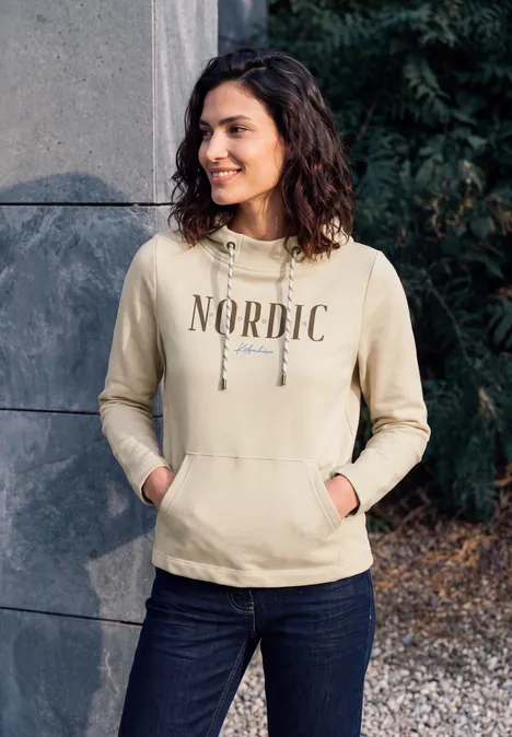 T-shirt à manches longues avec col cheminée et inscription pearl beige Eine Frau steht draußen vor einer Wand, trägt lächelnd einen hellbeigen Hoodie mit 'NORDIC'-Aufdruck.