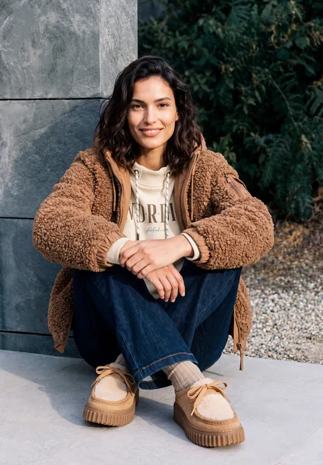 T-shirt à manches longues avec col cheminée et inscription pearl beige Junge Frau sitzt entspannt im Freien, trägt eine braune Teddyjacke, Hoodie und Jeans und lächelt in die Kamera.