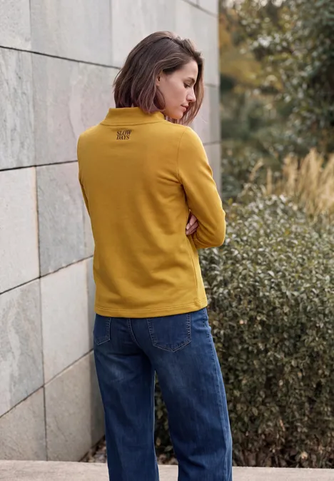T-shirt à manches longues avec col cheminée et inscription golden honey Frau in gelbem Pullover und Jeans lehnt an Wand, blickt zur Seite, umgeben von Sträuchern.