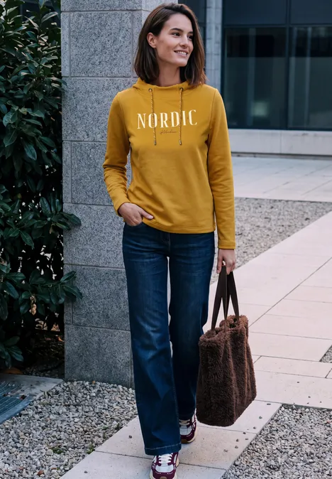 T-shirt à manches longues avec col cheminée et inscription golden honey Frau im senfgelben Pullover mit Schriftzug 'NORDIC', Jeans und Tasche steht lächelnd vor moderner Fassade.