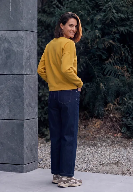 Shirt met lange mouwen, ronde hals en geribbelde structuur golden honey Eine Frau steht im Freien vor Sträuchern, blickt zurück und trägt ein senfgelbes Oberteil, blaue Jeans und Sneaker.