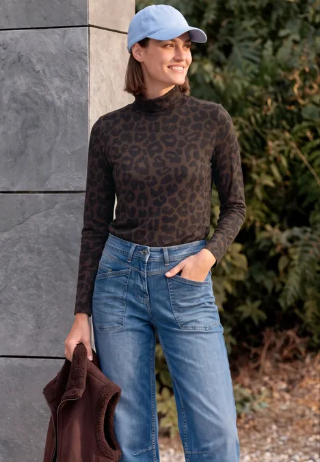 Shirt met lange mouwen, col en luipaardprint truffle brown Eine Frau lehnt lächelnd an einer Wand, trägt ein Leo-Rollkragen-Shirt, Jeans, hellblaue Kappe und hält eine Jacke.
