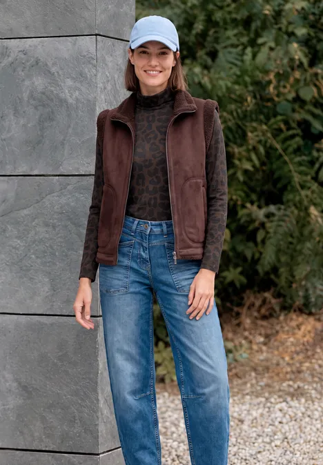 Shirt met lange mouwen, col en luipaardprint truffle brown Eine lächelnde Frau mit hellblauer Mütze, brauner Weste, schwarzem Pullover und Jeans vor Betonwand und Grün.