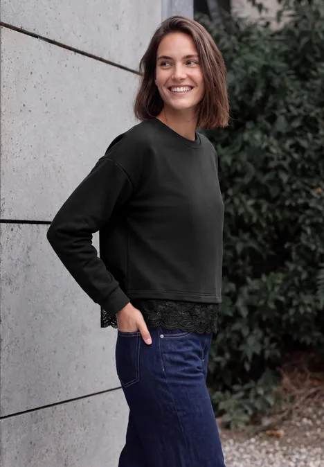 Eine lächelnde Frau in schwarzem Pullover und blauen Jeans lehnt an einer Wand.