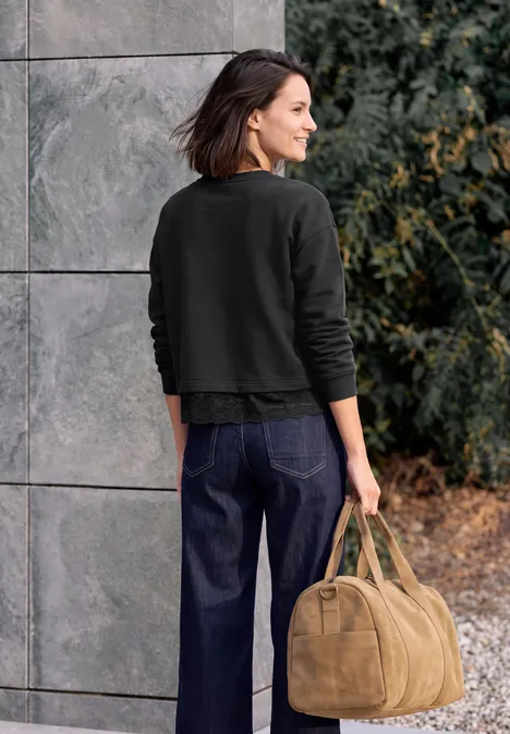 Frau von hinten in dunklem Pullover und Jeans, mit hellbrauner Tasche vor grauer Wand und Büschen.
