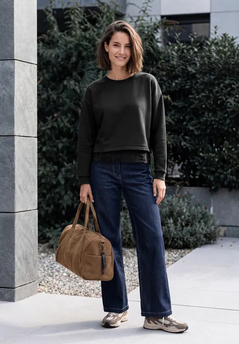 Frau in schwarzem Pullover, blauen Jeans und weißen Sneakern mit brauner Tasche steht vor Büschen.
