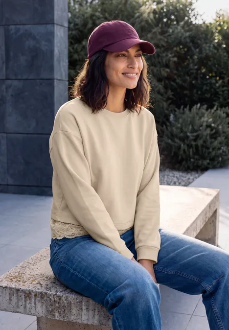 Frau mit Jeans, cremefarbigem Pullover und pinker Kappe sitzt lächelnd auf einer Steinbank.