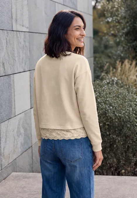 Eine lächelnde Frau in beigem Longsleeve mit Spitzenrand und Jeans lehnt an einer Steinmauer im Freien.