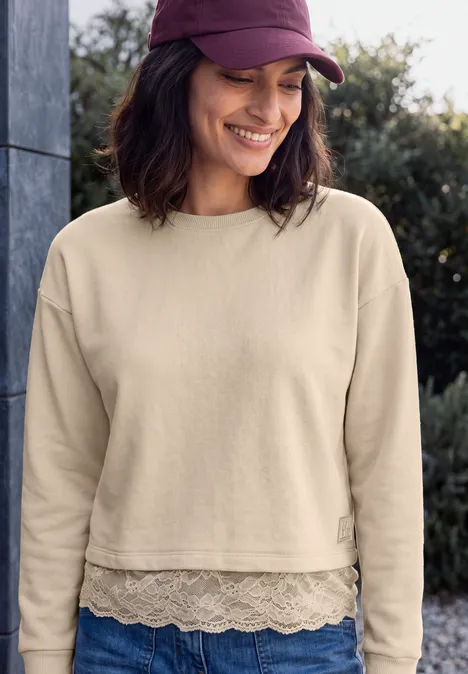 Eine lächelnde Frau in beigem cropped Sweatshirt mit Spitzenrand und violettem Cap.