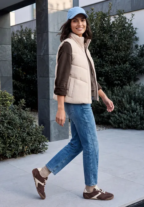 Shirt met lange mouwen, ronde hals en kanten rand truffle brown Vrouw in een beige bodywarmer, spijkerbroek en sneakers loopt glimlachend voor een gebouw met struiken.