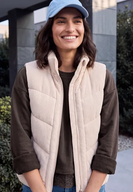 Shirt met lange mouwen, ronde hals en kanten rand truffle brown Glimlachende vrouw met lichtbeige doorgestikte bodywarmer, donker shirt en lichtblauwe pet voor modern gebouw.