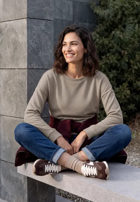 Shirt met lange mouwen en visgraatpatroon pearl beige Een vrouw zit buiten op een betonnen bank, lacht en draagt een trui, jeans en sneakers.