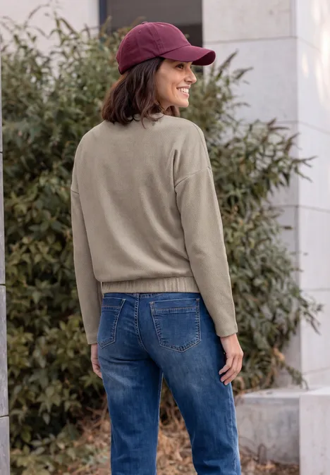 Shirt met lange mouwen en visgraatpatroon pearl beige Vrouw met rode pet, olijfgroene trui en spijkerbroek staat buiten en glimlacht over haar schouder.