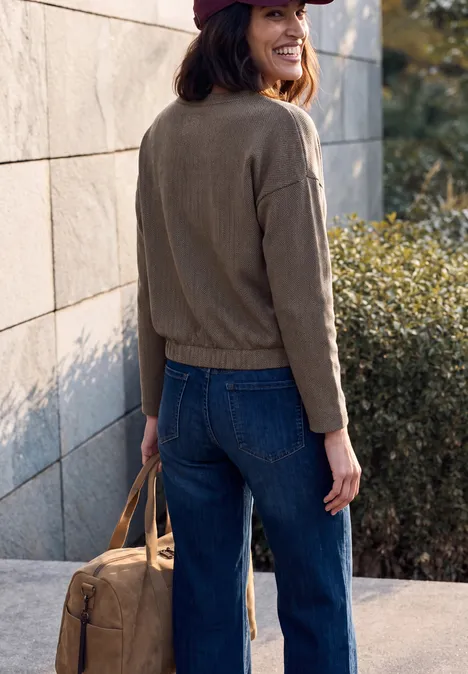 Langarm Shirt mit Herringbone-Muster truffle brown Frau im Jeans-Outfit, Jacke und Cap, trägt Tasche, steht an Wand im Freien.