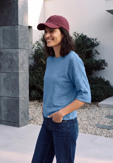 Femme souriante avec casquette rouge, t-shirt bleu et jean, debout devant un bâtiment moderne.