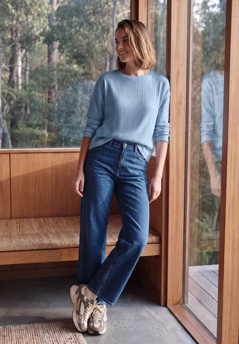 Femme en pull bleu et jean, debout près d'une fenêtre en bois, regardant vers la forêt depuis la maison.