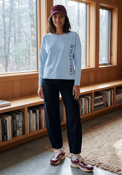 Vrouw in casual outfit (lichtblauw shirt, donkere broek, sneakers, pet) voor raam en boekenplank.