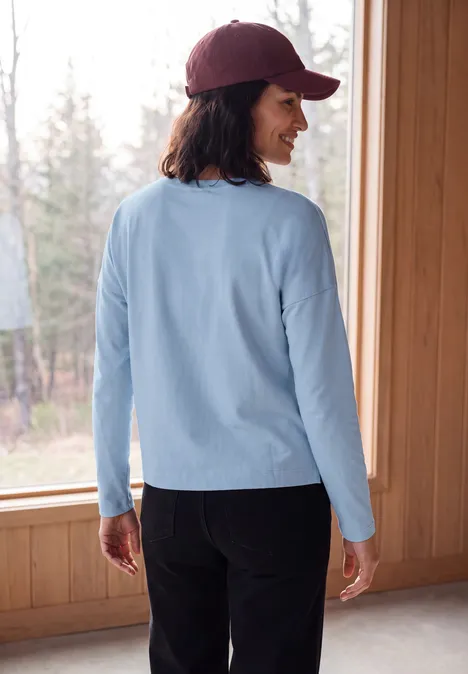 Vrouw van achteren met lichtblauw shirt, donkere broek en bordeaux pet, staand bij groot raam naar het bos.