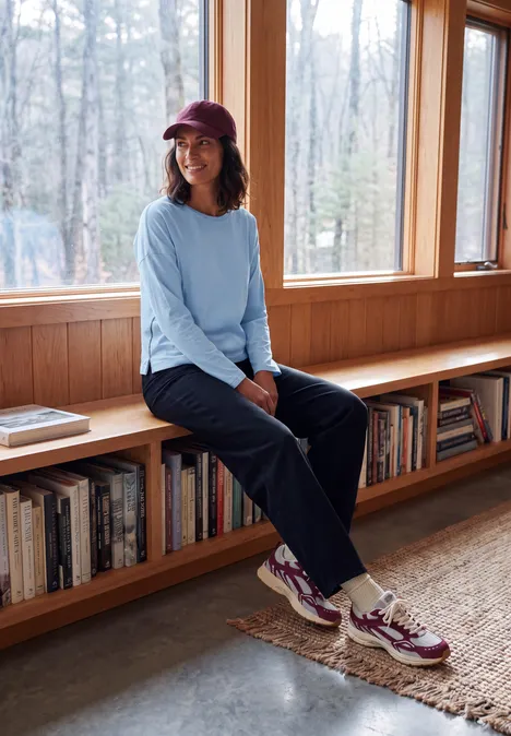 Vrouw met pet en sneakers zittend op een houten bank voor grote ramen met uitzicht op het bos.