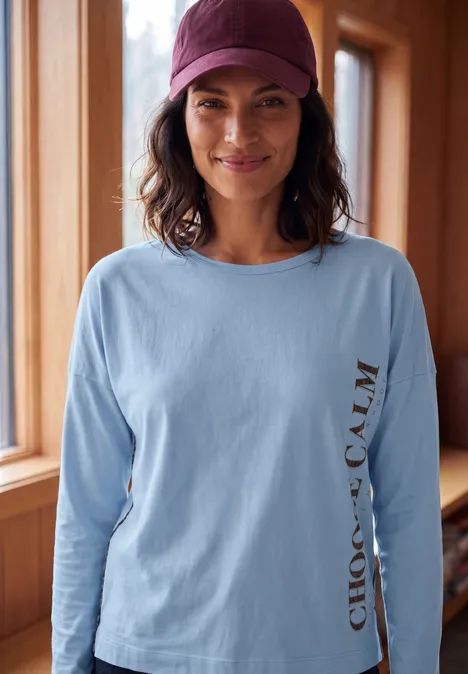 Vrouw glimlachend in lichtblauw shirt met 'CHOOSE CALM' en bordeaux pet bij een raam