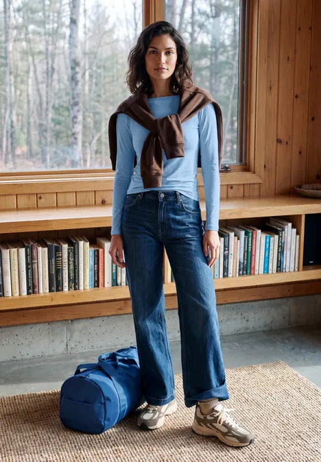 Effen shirt met lange mouwen en een boothals cosmopolitan blue Vrouw in licht trui, jeans en sneakers met reistas, staand voor boekenplank in houten kamer.