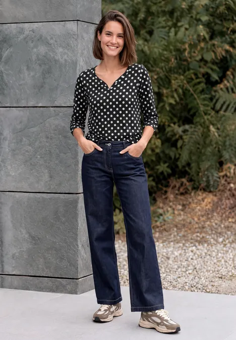 Frau in schwarzer Bluse mit weißen Punkten, weitem dunkelblauen Jeans, Sneakern, lächelnd vor Steinwand und Bäumen.