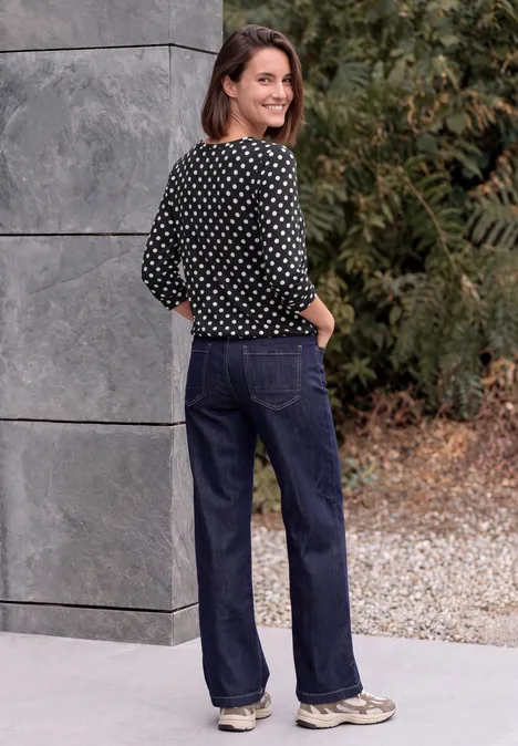 Eine Frau in gepunkteter Bluse und weiter Jeans steht lächelnd vor einer grauen Mauer im Freien.