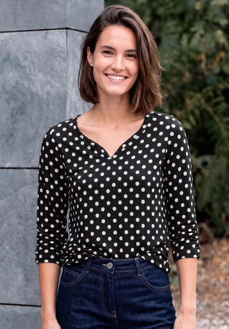 Eine lächelnde Frau in gepunkteter Bluse und Jeans steht vor einer Mauer und Pflanzen.