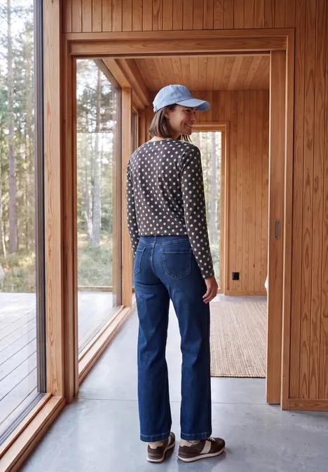 Person mit blauer Mütze, gepunktetem Pullover und Jeans steht in hellem Holzhaus, Wald draußen sichtbar.