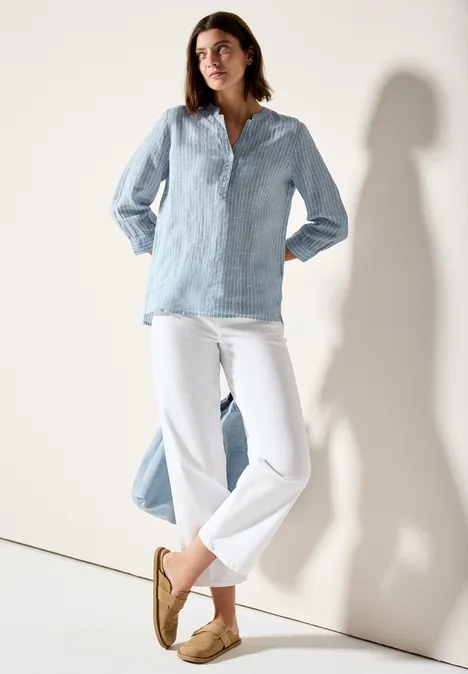 Vrouw met lichtblauwe blouse, witte broek en beige schoenen leunt nonchalant tegen een muur.