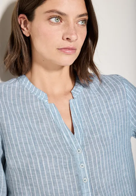 Vrouw met bruin haar draagt een lichtblauw gestreepte blouse en kijkt opzij.