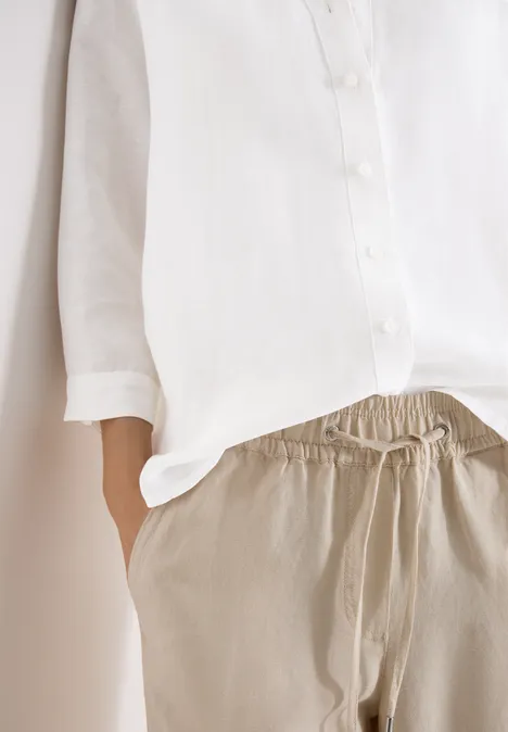 Eine Person trägt eine weiße Bluse und bequeme beige Hose, Hand in der Hosentasche.